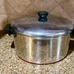 Pode incluir: Uma panela de aço inoxidável com tampa e alças pretas. A panela tem fundo cor de cobre e um botão azul escuro na tampa. A panela está sobre uma bancada salpicada.