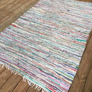 Pu&ograve; includere: Un tappeto rettangolare multicolore di stracci con un motivo a righe. Il tappeto presenta un mix di colori tra cui bianco, rosso, blu e verde. Il tappeto ha un bordo sfrangiato ed &egrave; posizionato su un terrazzo di legno.