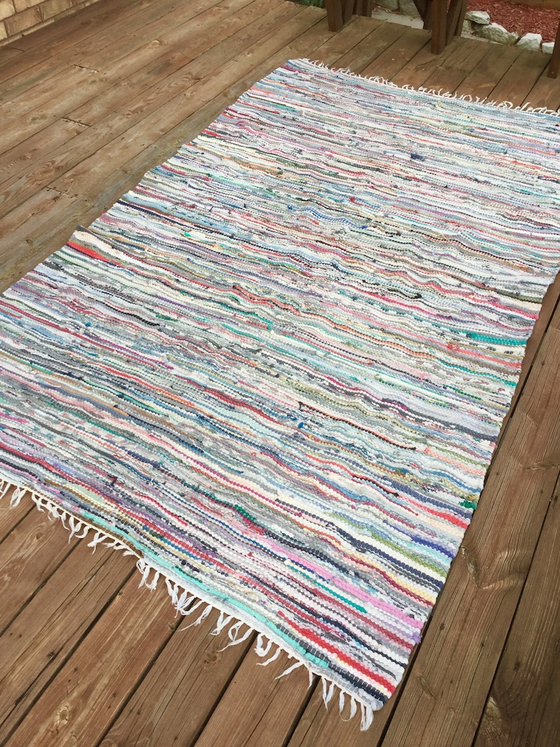 Pu&ograve; includere: Un tappeto rettangolare multicolore di stracci con un motivo a righe. Il tappeto presenta un mix di colori tra cui bianco, rosso, blu e verde. Il tappeto ha un bordo sfrangiato ed &egrave; posizionato su un terrazzo di legno.