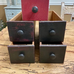May include: A wooden storage unit with five drawers arranged in a tiered formation. The top drawer is painted red, while the four lower drawers are painted black with distressed edges. Each drawer features a small, round, dark-colored wooden knob.