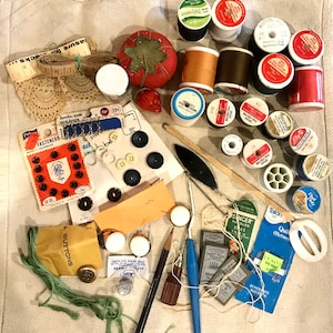 May include: A collection of sewing supplies including spools of thread in various colors, buttons, needles, and a thimble. There are also some small packages of fasteners and a ruler.