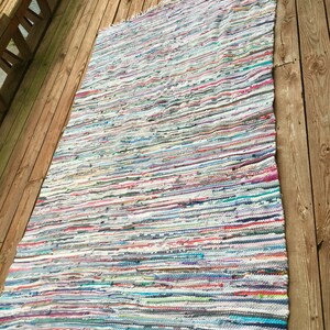Pu&ograve; includere: Un tappeto rettangolare multicolore con frange su un bordo. Il tappeto presenta strisce orizzontali in vari colori, tra cui bianco, blu, rosa e verde. Il tappeto &egrave; posizionato su un terrazzo di legno.
