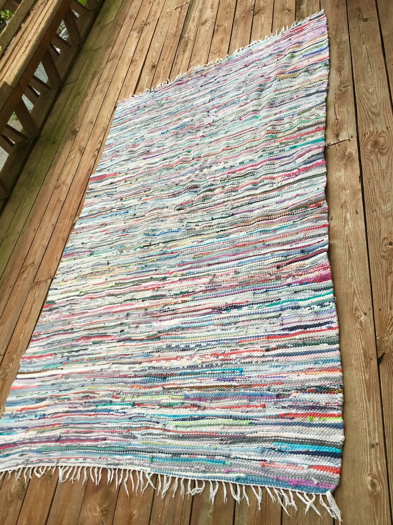 Pu&ograve; includere: Un tappeto rettangolare multicolore con frange su un bordo. Il tappeto presenta strisce orizzontali in vari colori, tra cui bianco, blu, rosa e verde. Il tappeto &egrave; posizionato su un terrazzo di legno.