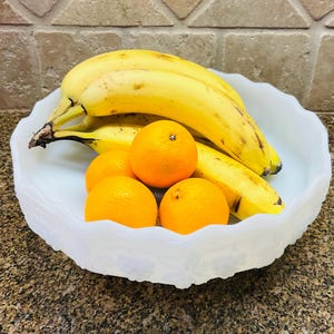 Op de afbeelding: Een witte keramische kom met een decoratief ontwerp, gevuld met gele bananen en oranje fruit.