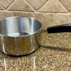 Op de afbeelding: Een roestvrijstalen steelpan met een zwarte handgreep. De pan is rond met een gebogen lichaam en een enkele, lange handgreep. De handgreep is zwart en heeft een gebogen vorm. De pan staat op een gespikkeld aanrecht.