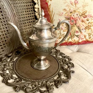 May include: A silver-toned metal teapot featuring a tapered body, a curved spout, a handle with ornate detailing, and a matching lid with a finial. The teapot rests on a circular, dark-colored decorative base with an intricate, scrolled border.