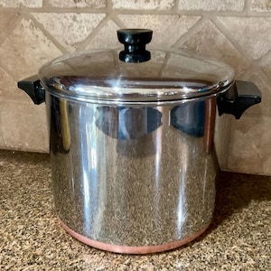 Puede incluir: Una olla de cocina de acero inoxidable con tapa y asas negras. La olla tiene una base de color cobre y un pomo negro en la tapa. La olla está sobre una encimera moteada.