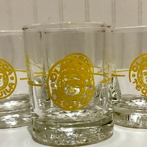 May include: Three clear glass shot glasses with a yellow Olmeca Tequila logo. Each glass has a gold rim and a gold band around the middle.