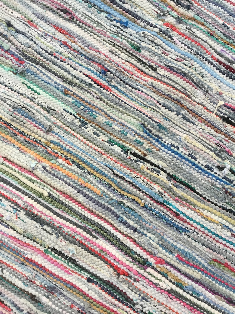 Pu&ograve; includere: Primo piano di un tappeto tessuto multicolore. Il tappeto presenta un motivo a strisce diagonali in vari colori, tra cui grigio, bianco, rosso, blu e verde. La trama sembra essere fatta di strisce di tessuto.