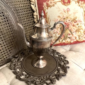 May include: A silver-toned metal coffee pot with a tall, tapered body, a curved handle, and a pointed lid finial. The pot features a decorative rope-like band around the midsection and rests on a circular pedestal base with a smooth, reflective finish.