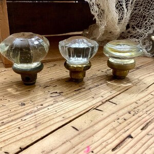 May include: Three clear glass knobs with faceted surfaces and brass-toned metal bases. Each knob features a distinct shape, ranging from a larger rounded design to smaller, flatter profiles, all mounted on circular metallic hardware components.