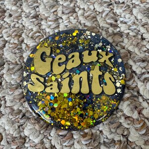 May include: A round, black resin coaster with gold glitter and the words "Geaux Saints" in gold lettering.