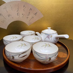 Op de afbeelding: Een wit theeservies met een bloemenmotief op een houten dienblad. De set bevat een theepot en vijf kopjes.