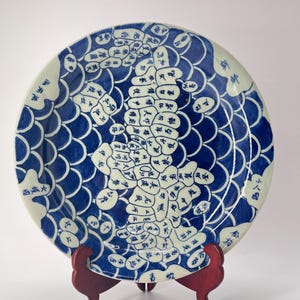May include: A blue and white porcelain plate with a map of Japan in Japanese characters. The plate is decorated with a blue and white fish scale pattern. The plate is on a wooden stand.