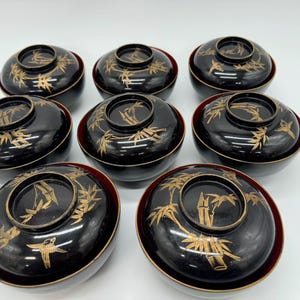 May include: A collection of eight black lacquer bowls with lids. Each bowl features a gold bamboo design on the lid and a red interior. The bowls are arranged in a circular pattern.