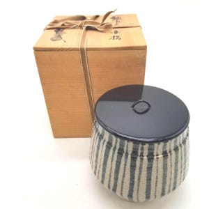 May include: A ceramic tea container with a black and white vertical striped pattern and a glossy black lid, accompanied by a light brown wooden storage box tied with a fabric ribbon. The wooden box features black Japanese calligraphy on its lid.