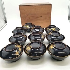 May include: A set of ten black lacquer bowls with matching lids, each featuring gold painted botanical and geometric motifs. The bowls are arranged in rows in front of a rectangular wooden storage box with a smooth, light brown grain finish.