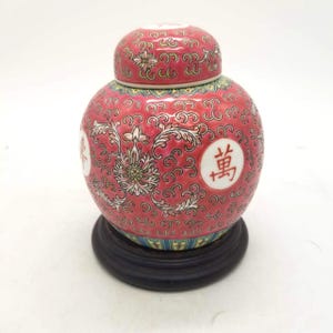 Könnte beinhalten: Ein rot-weißes Keramik-Ingwerglas mit Deckel, mit floralen Mustern und chinesischen Schriftzeichen. Das Glas steht auf einem dunklen Holzsockel. Das Glas ist ein dekorativer Gegenstand.