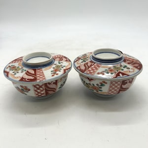 Puede incluir: Dos cuencos de cerámica a juego con tapas. Los cuencos tienen una base blanca con patrones florales y geométricos rojos, verdes y dorados. Las tapas tienen un anillo central elevado y están decoradas con patrones similares. Los bordes están adornados con azul y blanco.