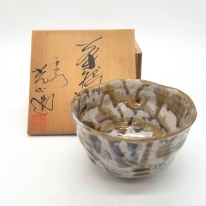 Pode incluir: Tigela de cerâmica artesanal com uma superfície texturizada única em tons de castanho, branco e azul. A tigela tem uma borda irregular e está em frente a uma caixa de madeira com caligrafia japonesa. Uma obra de arte funcional.