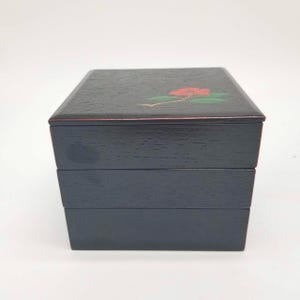 May include: A black, three-tiered bento box with a floral design on the lid. The flower is red with green leaves. The box has a rectangular shape and a red trim.