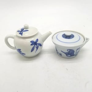 May include: A white ceramic teapot and covered bowl set. The teapot has a curved handle and spout, with blue floral designs. The bowl has a lid and a blue design around the rim and on the side. The set is likely for tea service.