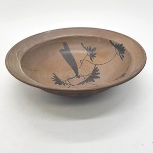 Könnte beinhalten: Eine braune Keramikschale mit breitem Rand und flachem Innenraum. Die Schale hat ein schwarzes Blumendesign auf der Innenseite. Die Schale ist rund und scheint handgefertigt zu sein.