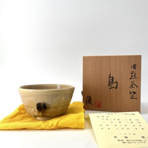 Puede incluir: Un cuenco de cerámica beige con una mancha oscura, sobre un paño amarillo. Una caja marrón con caracteres japoneses y un papel con texto están detrás del cuenco. El cuenco tiene un aspecto rústico y artesanal.