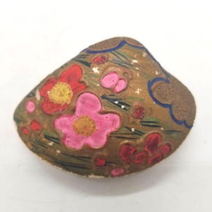 May include: A decorative seashell painted with a gold background featuring hand-painted red and pink flowers with yellow centres, green grass-like blades, and blue curved line accents. The shell has a textured surface and a natural, irregular shape.