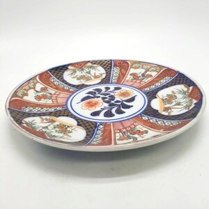 Puede incluir: Un plato decorativo ovalado con un diseño floral y paisajístico detallado. El plato presenta un motivo floral central en naranja y azul, rodeado de paneles alternos rojos, azules y blancos con detalles dorados. El plato es probablemente de cerámica.