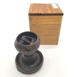 May include: A dark, metallic, circular object with a central handle and a wide, flat base, positioned in front of a rectangular wooden box with a lid. The box features a light brown wood grain and a small white rectangular label on the top surface.