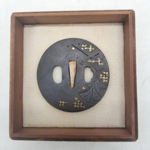 Op de afbeelding: Een cirkelvormige zwaardstootplaat van donker metaal, of tsuba, voorzien van goudkleurige ingelegde stervormige patronen en een bladmotief. Het object heeft een centrale langwerpige opening en twee zijopeningen, geplaatst in een vierkante houten doos.