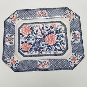 Könnte beinhalten: Rechteckige Keramik-Servierplatte mit Blumen- und Vogeldesign. Die Mitte zeigt rosa Rosen und einen blauen Vogel inmitten blauer Blätter. Der Rand hat ein blau-weißes geometrisches Muster mit rosa und roten floralen Akzenten.