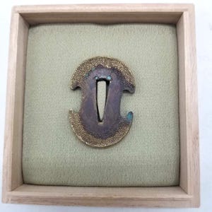 Peut inclure: Un tsuba japonais, garde d'épée, présenté dans une boîte en bois. Le tsuba est de couleur bronze foncé avec un motif texturé et orné et une ouverture centrale. La boîte a un intérieur en tissu vert clair.