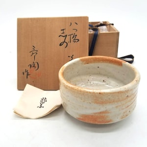May include: A ceramic tea bowl with a cream-coloured interior and a reddish-brown rim and accents. The bowl is accompanied by a wooden box with Japanese calligraphy and a small paper with Japanese text. A blue ribbon is tied around the box.
