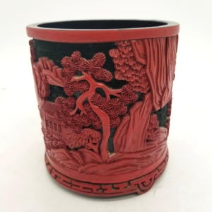 Op de afbeelding: Een rode, gebeeldhouwde pennenhouder met een zwart interieur. De buitenkant heeft een gedetailleerd reliëf van een landschap met bomen, gebouwen en bergen. De pennenhouder heeft een decoratieve basis en rand.