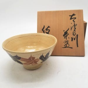 May include: A beige ceramic bowl with a footed base, decorated with brown and dark blue leaf designs. The bowl is paired with a wooden box featuring Japanese calligraphy. The bowl has a diameter of approximately 6 inches.