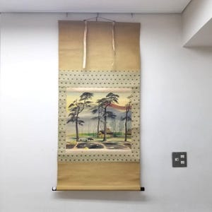 Peut inclure: Peinture japonaise sur rouleau représentant un paysage avec des arbres, un village et des montagnes. L'œuvre est encadrée d'une bordure à pois et suspendue à une tringle métallique avec des rubans beiges.