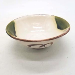 May include: A shallow ceramic bowl with a cream-coloured interior and exterior. The rim features a thin brown painted line, while two dark green patches are painted on opposite sides. A brown swirling graphic is painted on the front exterior.