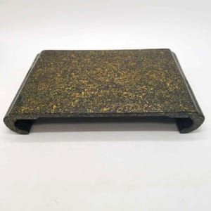 May include: A rectangular, dark-colored tray with a speckled, textured surface in shades of black and gold. The tray has a curved edge on each end, and is elevated slightly off the surface.