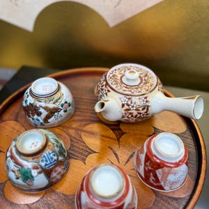 Puede incluir: Un juego de cuatro pequeñas tazas de té blancas con diseños florales y geométricos, y una tetera a juego con un diseño floral, todo en una bandeja de madera.
