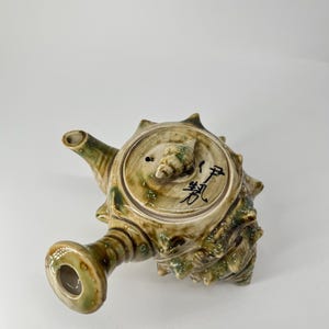 May include: A ceramic teapot with a unique, textured design resembling a sea shell. The teapot is primarily olive green and brown with a spout, handle, and lid. The lid has a small knob and the teapot has Japanese characters on the lid.