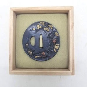 Op de afbeelding: Een cirkelvormige zwaardstootplaat van donker metaal, of tsuba, met gedetailleerde reliëfs van een figuur met een bezem en een boom met goudkleurige bladeren. Het stuk wordt gepresenteerd in een vierkante houten doos met een lichtgroene stoffen voering.