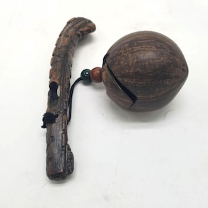 May include: A handcrafted wooden rattle with a dark brown finish. The rattle features a textured handle and a round, hollow bell with a split. A black cord and beads connect the bell to the handle. The overall design is rustic and natural.