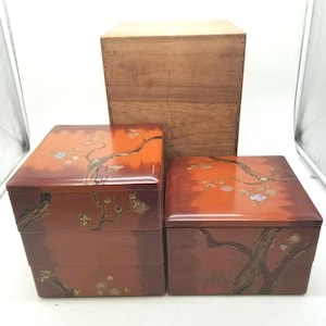 May include: A set of two square, tiered lacquer boxes in a deep reddish-brown finish, featuring inlaid mother-of-pearl floral branch designs. The boxes are accompanied by a plain, light-colored wooden storage container positioned behind them.