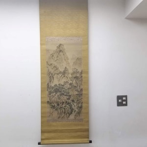 May include: A vertical, antique scroll painting depicting a mountainous landscape with trees and foliage. The artwork features muted tones of green, brown, and gray, with a gold-colored border and a black rod at the bottom for hanging.