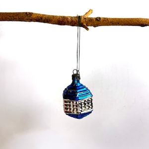 Peut inclure: Une décoration de Noël vintage, bleue et argentée, suspendue à une branche brune. L'ornement présente un motif géométrique avec une bande argentée et un dessus bleu. Le fond est blanc uni.