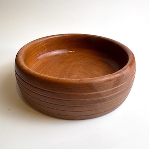 Puede incluir: Un cuenco redondo de madera con un acabado liso y pulido de color marrón cálido. El exterior del cuenco presenta una serie de cinco ranuras horizontales y paralelas talladas en el lateral, creando un aspecto texturizado alrededor de la base.