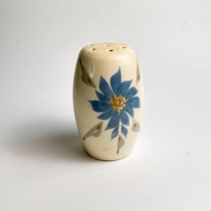 Puede incluir: Salero de cerámica color crema con un diseño floral azul. La flor tiene un centro dorado y pétalos azules. El salero tiene agujeros en la parte superior para dispensar sal y está decorado con hojas grises.