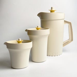 Può includere: Un set di tre brocche e contenitori color crema. La brocca più grande ha un manico e un coperchio con la parte superiore gialla. Gli altri due contenitori hanno anche coperchi con la parte superiore gialla. Il set è in materiale opaco, su sfondo bianco.